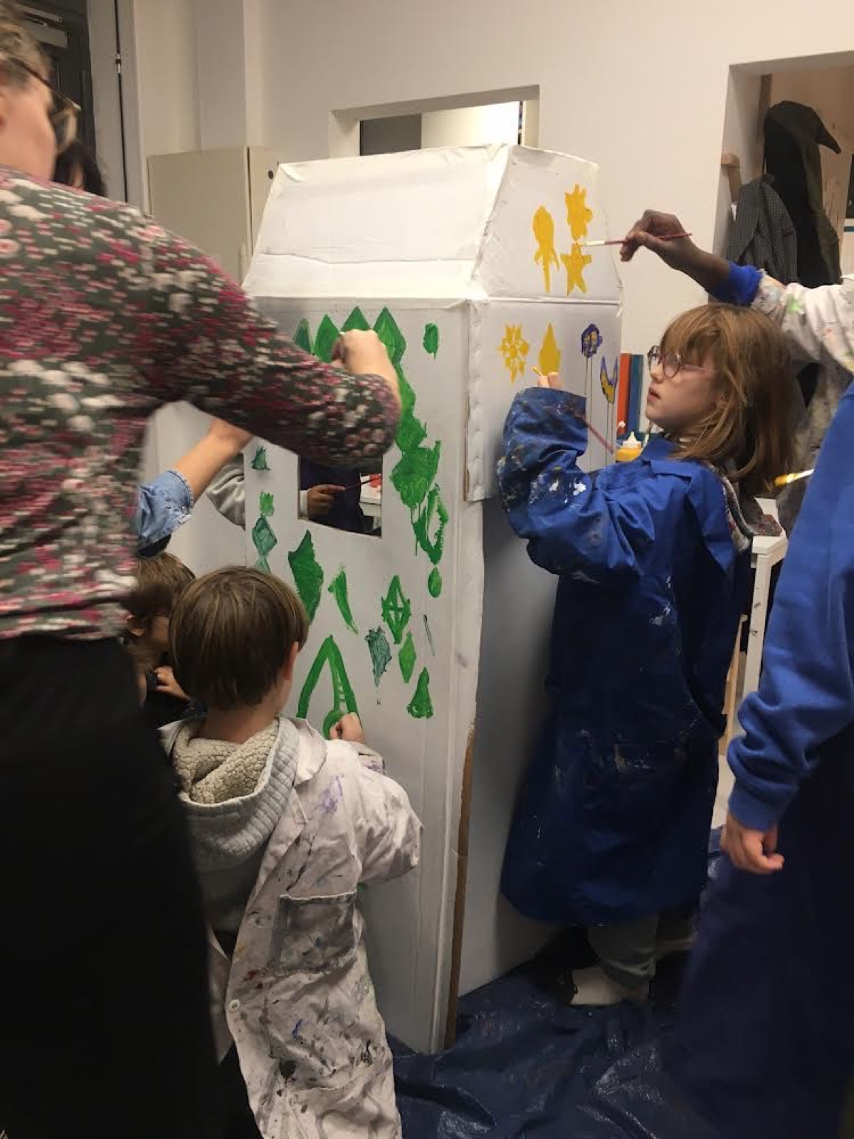 Des enfants peignent une cabane en carton avec un adulte
